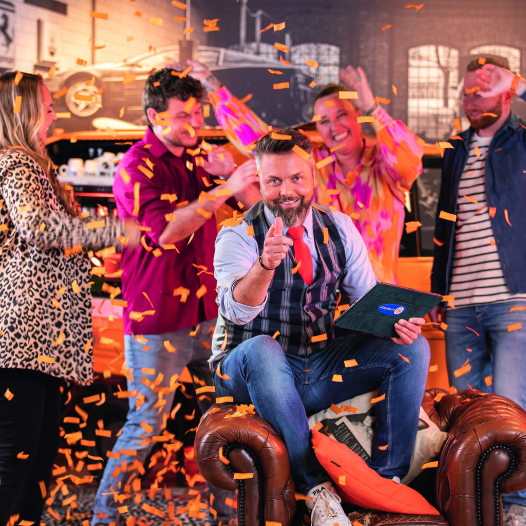 Op de afbeelding zie je een man omringd door blije mensen en oranje confetti.